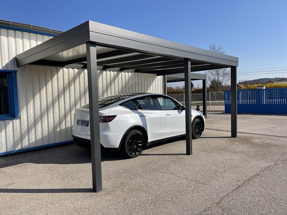 Une berline blanche est garée sous un abri de voiture gris, à côté d'un bâtiment beige, avec une clôture bleue en arrière-plan.