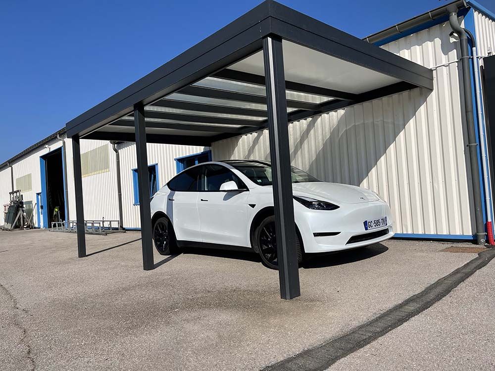 Voiture blanche garée sous un abri métallique à côté d'un bâtiment industriel beige par une journée ensoleillée