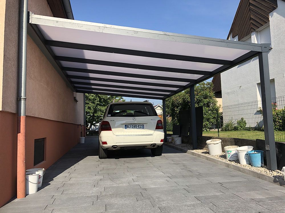 Un SUV blanc est garé sous un abri métallique à côté d'une maison, avec des seaux le long de l'allée.