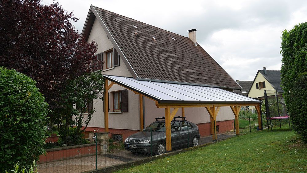 Maison au toit de tuiles pentu et abri voiture à côté d'une voiture garée, dans une cour suburbaine herbeuse sous un ciel nuageux.