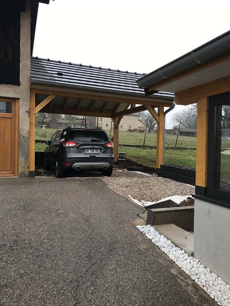 Voiture garée sous un abri en bois à côté d'une maison moderne, sur une allée de gravier.