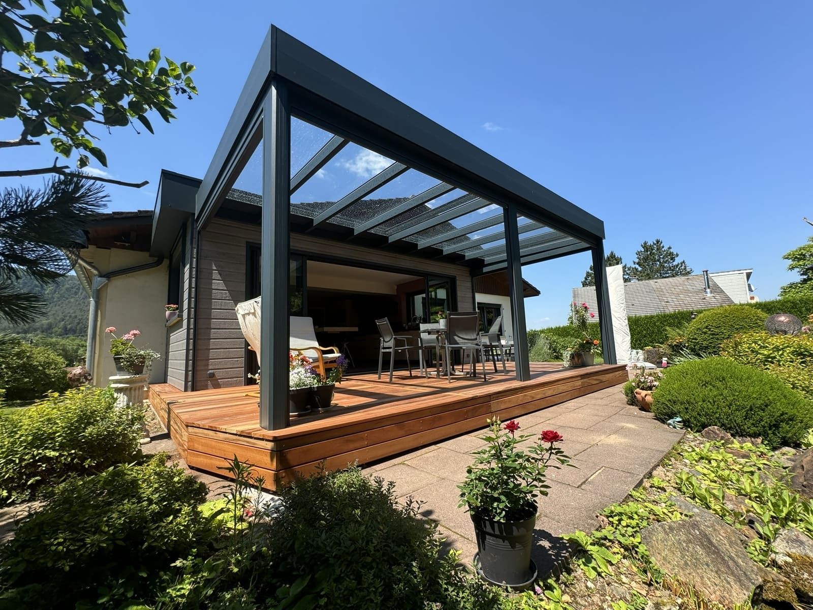 Patio moderne avec pergola noire, terrasse en bois, coin salon extérieur et jardin sous un ciel bleu