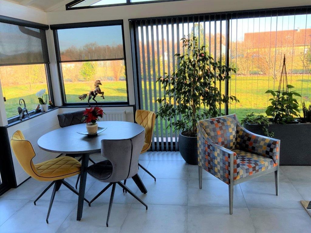 Véranda moderne avec table ronde, chaises colorées, grandes plantes et baies vitrées donnant sur un champ
