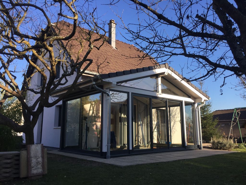 Petite maison blanche avec un toit rouge et une véranda vitrée dans une cour ensoleillée