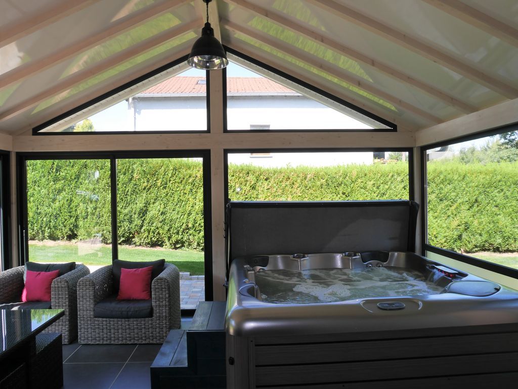 Terrasse couverte avec sièges en osier et bain à remous, donnant sur une haie verdoyante et un jardin.