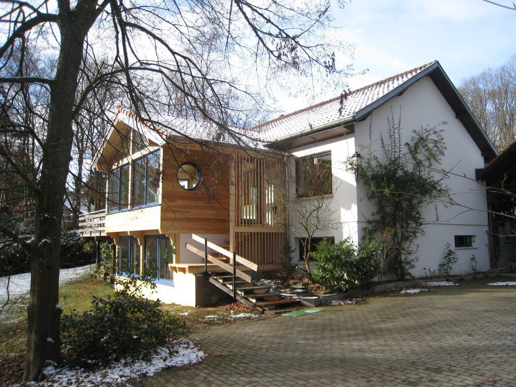 Maison à deux étages avec porche et escalier en bois, située à côté d'arbres dénudés et d'une allée pavée légèrement enneigée.