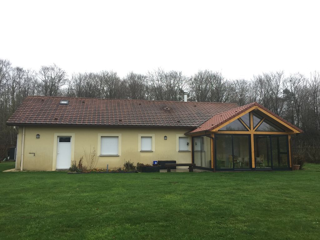 Maison jaune de plain-pied avec un toit marron et une véranda vitrée, située dans un jardin gazonné.