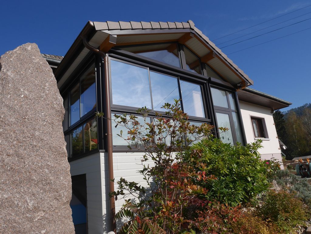 Maison moderne avec de grandes fenêtres réfléchissantes et un jardin, vue depuis un mur de pierre sous un ciel dégagé.