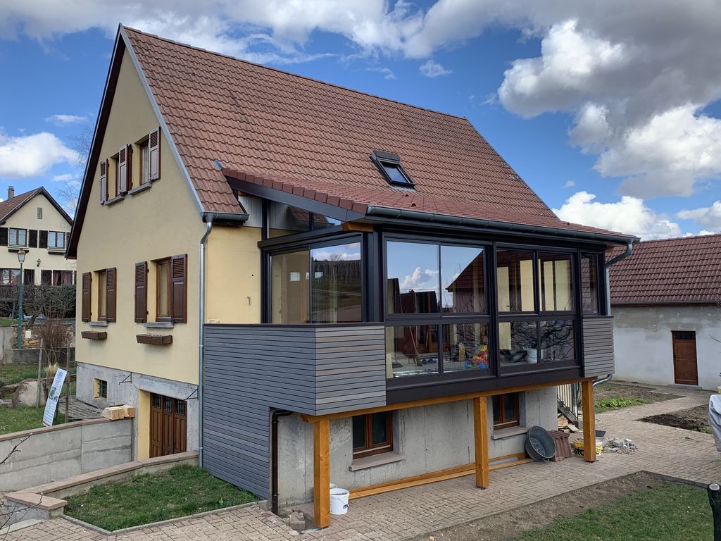 Maison moderne à deux étages avec toit rouge, balcon vitré et façade inférieure grise sous un ciel bleu