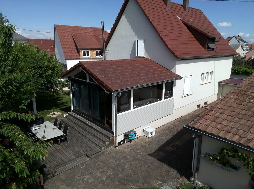 Maison blanche au toit et au porche rouges, vue d'en haut, entourée d'arbres et de maisons voisines.