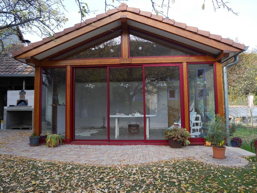 Petite véranda vitrée à cadre rouge et toit pointu, entourée d'une cour recouverte de feuilles.