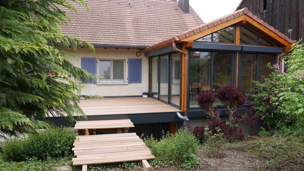 Extérieur de la maison avec terrasse et rampe en bois, véranda vitrée et jardin luxuriant.