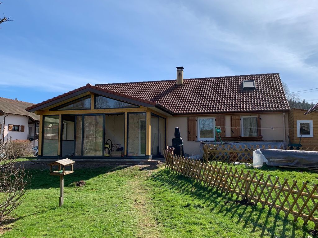 Maison de plain-pied avec toit de tuiles rouges, grandes fenêtres et jardin gazonné sous un ciel bleu.