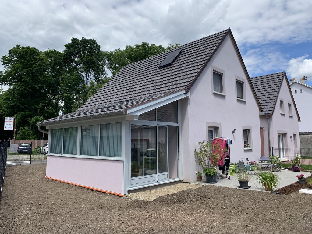 Maison blanche moderne avec toit gris foncé, véranda vitrée, cour gravillonnée et fleurs en pot sous un ciel nuageux