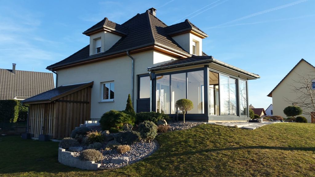 Maison blanche moderne avec toit foncé, véranda vitrée et jardin paysager à l'avant, baigné de lumière naturelle.