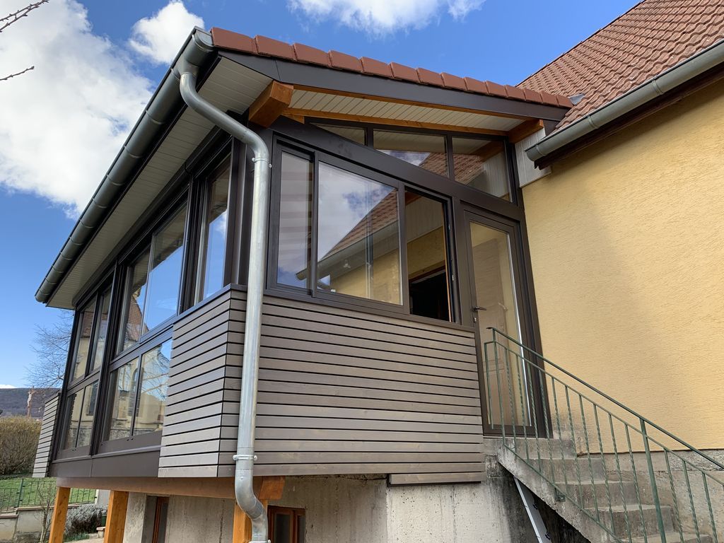 Véranda fermée avec de grandes fenêtres et un revêtement métallique, attenante à la maison et accessible par un escalier extérieur.
