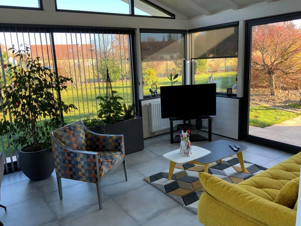 Véranda moderne avec chaises colorées, canapé jaune, télévision et grandes fenêtres donnant sur un jardin.