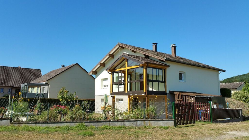 Maison de banlieue avec une véranda vitrée, des murs blancs et un jardin sous un ciel bleu azur.