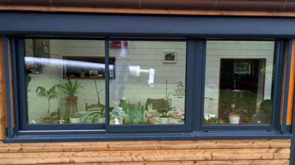 Extérieur d'une maison avec une large fenêtre bleu foncé, laissant apparaître des plantes en pot à l'intérieur et un bardage en bois en dessous.
