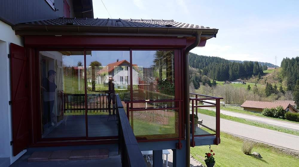 Porche d'une maison rouge avec rambarde donnant sur une colline verdoyante et la route