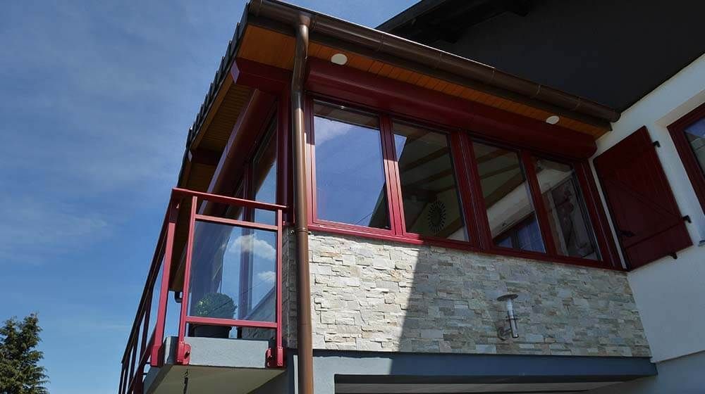 Extérieur d'une maison moderne avec fenêtres à cadre rouge, mur en pierre et toit en surplomb sous un ciel bleu