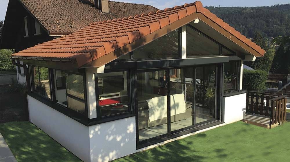 Véranda moderne en verre avec toit de tuiles rouges et murs blancs dans un jardin verdoyant