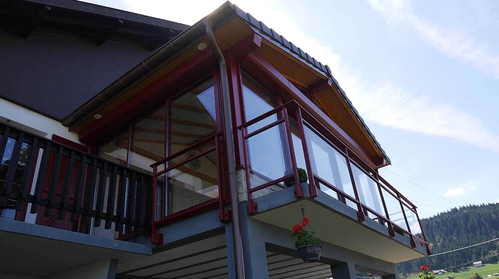 Balcon d'une maison moderne avec rambardes rouges et panneaux de verre sous un toit en pente, donnant sur une colline verdoyante.