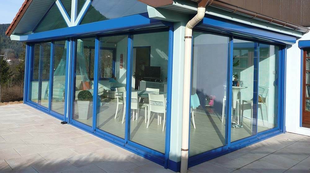Véranda vitrée à cadre bleu, attenante à un bâtiment, avec des chaises blanches à l'intérieur et une terrasse carrelée à l'extérieur.