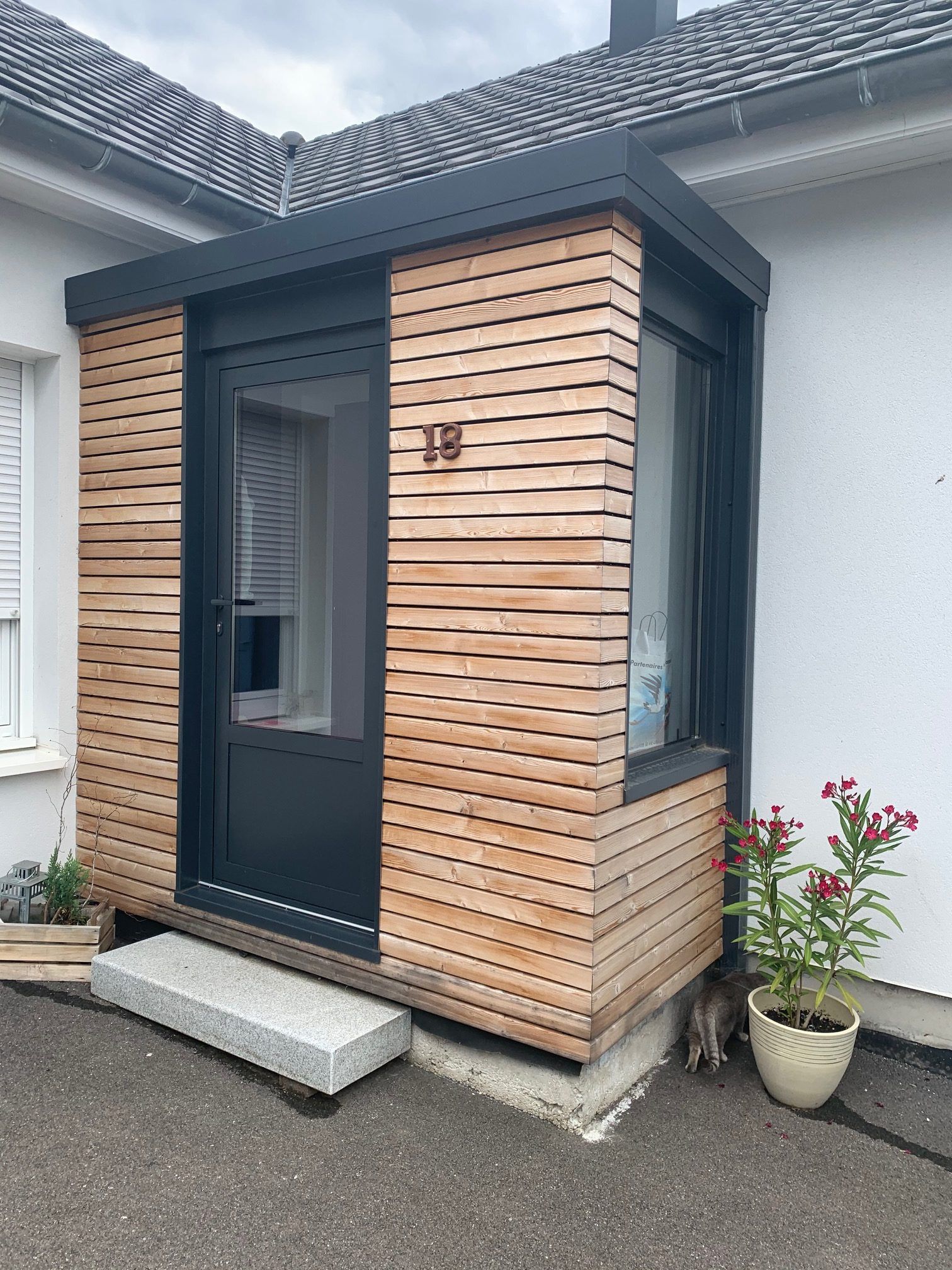 Petit porche d'entrée moderne avec bardage en lattes de bois, porte foncée et marche en béton à côté d'une maison