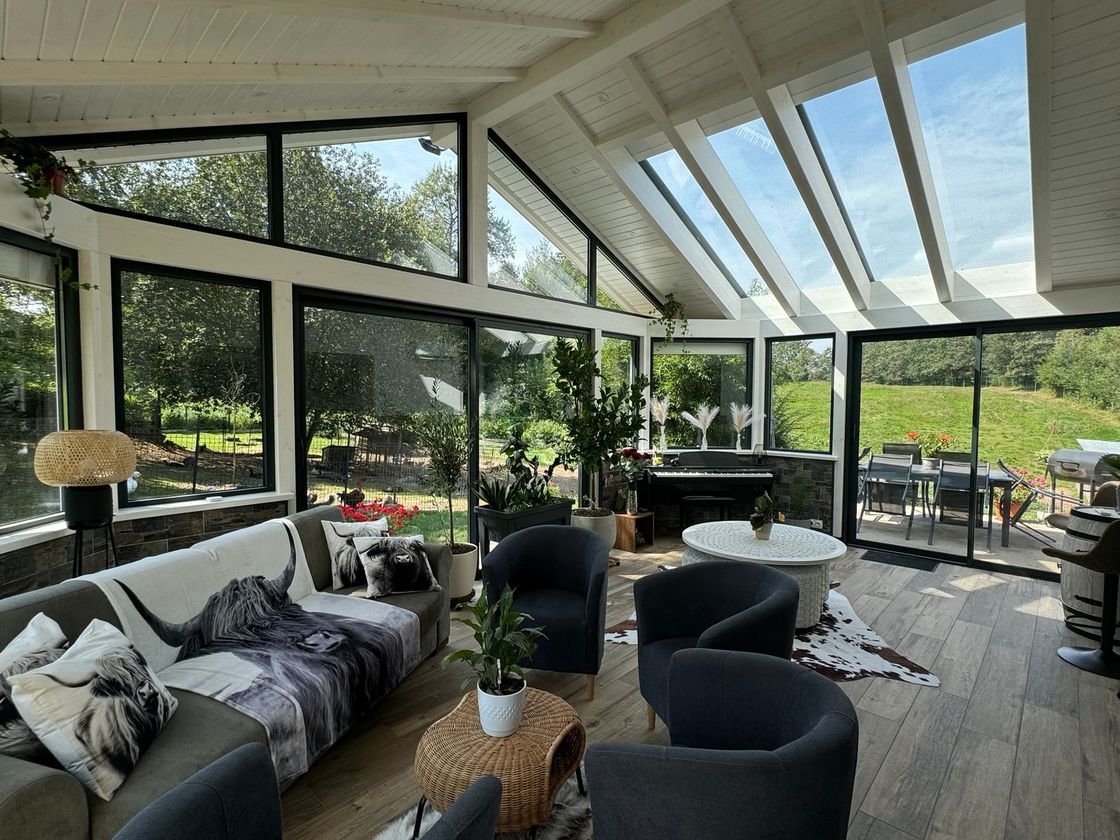 Véranda lumineuse avec parois vitrées, canapé gris, chaises noires et piano donnant sur une terrasse verdoyante