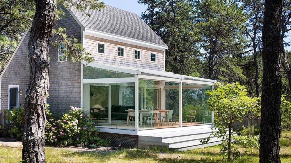 Maison moderne avec véranda vitrée et terrasse en bois entourée d'arbres et de verdure.