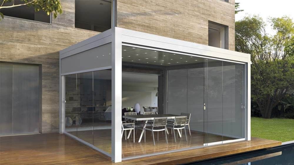 Véranda moderne aux parois de verre, attenante à la maison, avec table et chaises sur une terrasse en bois.
