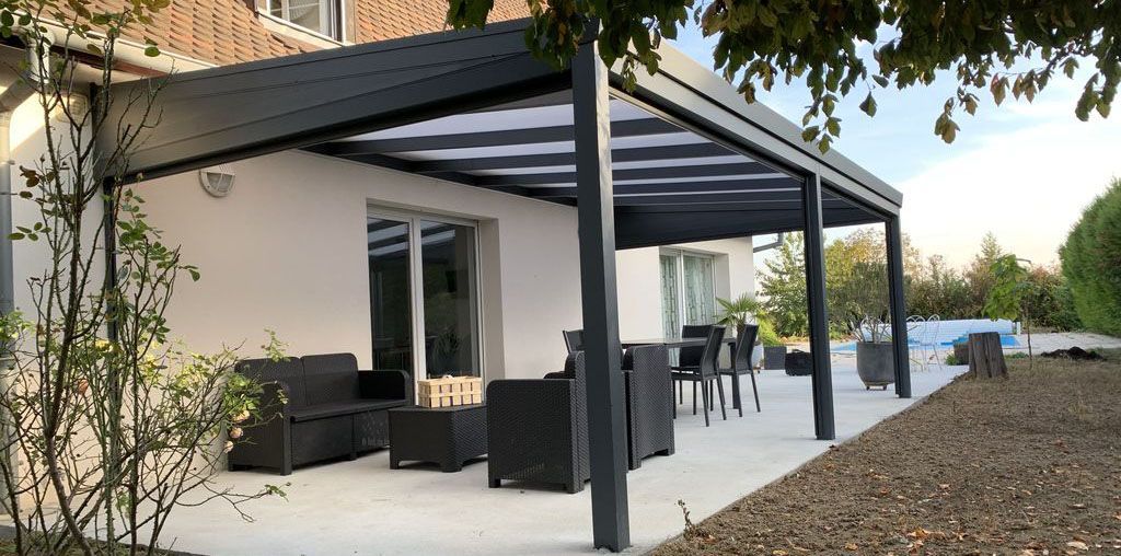 Patio moderne avec mobilier noir sous une pergola grise, à côté d'une maison, donnant sur une cour gravillonnée et une piscine.