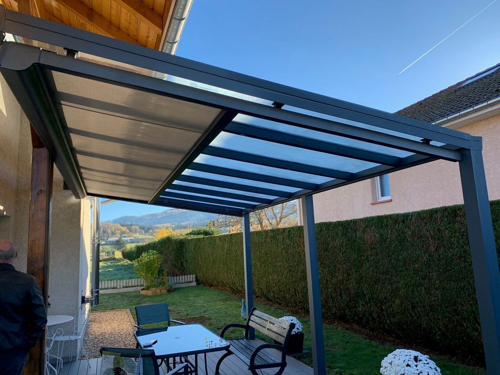 Terrasse avec pergola métallique, table en verre, banc, haie et mur de maison dans un jardin arrière