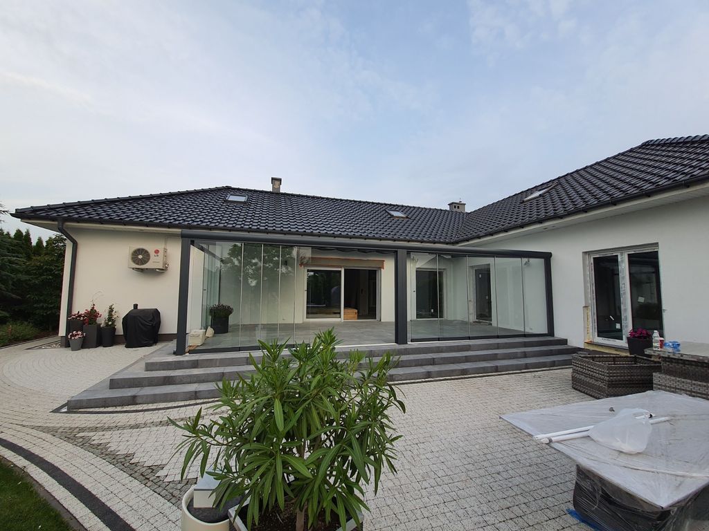 Maison moderne de plain-pied avec toit de tuiles grises, grandes fenêtres et patio pavé sous un ciel nuageux