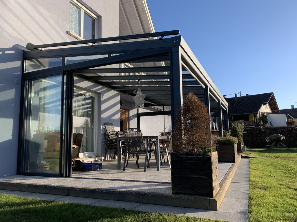 Terrasse vitrée ensoleillée attenante à la maison, avec mobilier de jardin et pelouse verte.