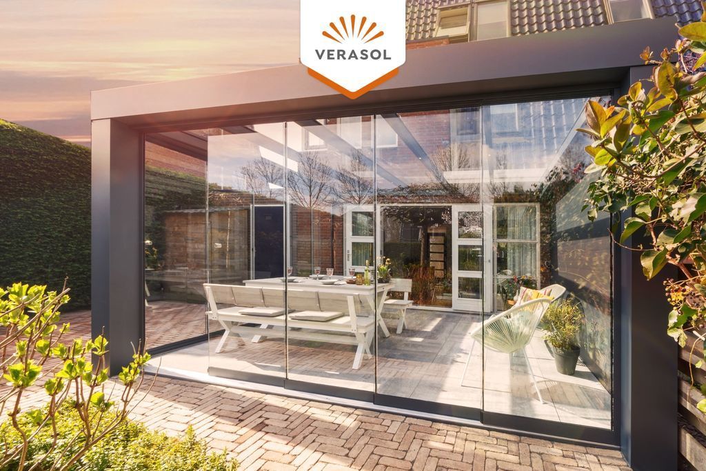 Terrasse moderne en verre avec table à manger blanche, encadrée de verdure et baignée de soleil.