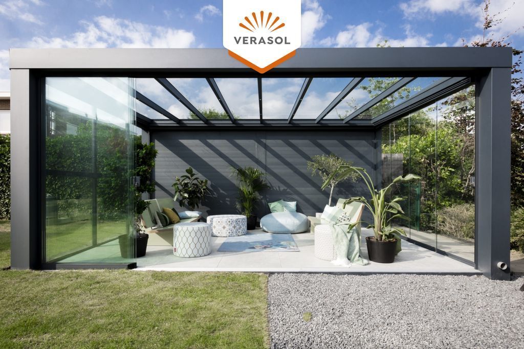 Pergola moderne en verre avec structure noire et coin salon extérieur confortable sur une terrasse en bordure de pelouse