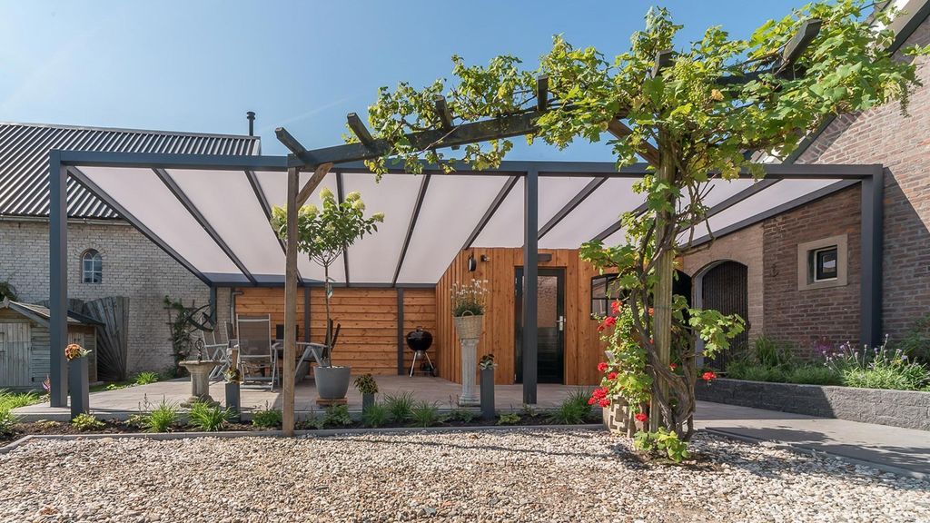 Cour intérieure moderne avec pergola, vignes, sol en gravier et bâtiments en pierre et en bois sous un ciel bleu azur.