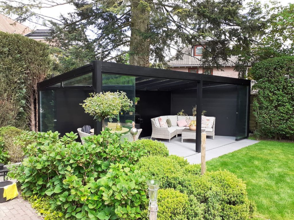 Pergola de jardin moderne noire avec sièges blancs sur une terrasse, entourée d'arbustes et d'une pelouse verdoyante.
