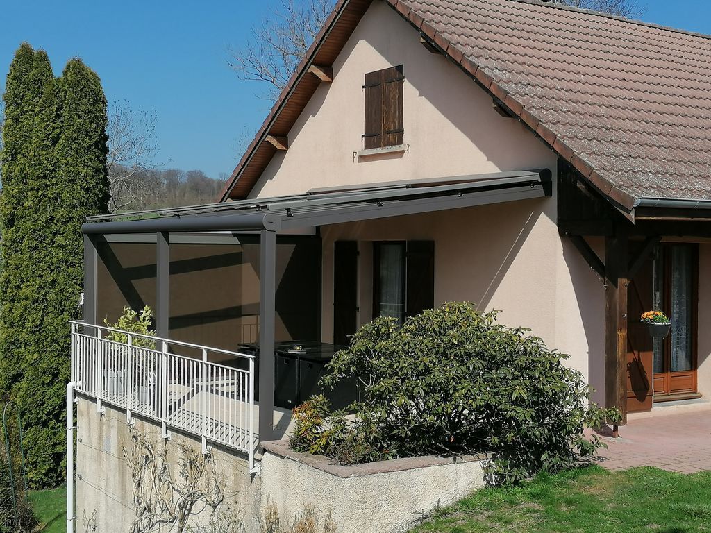 Petite maison beige avec un porche couvert, une balustrade blanche et un arbuste vert devant.