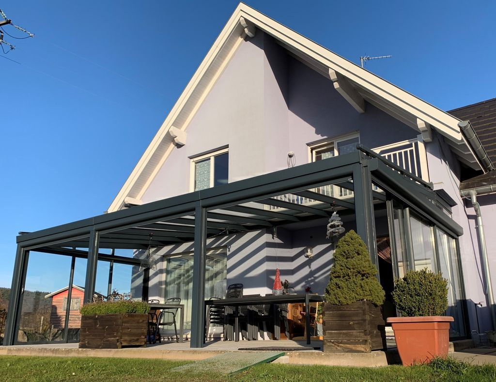 Maison moderne avec une véranda vitrée, du mobilier de jardin et des plantes en pot sous un ciel bleu azur.