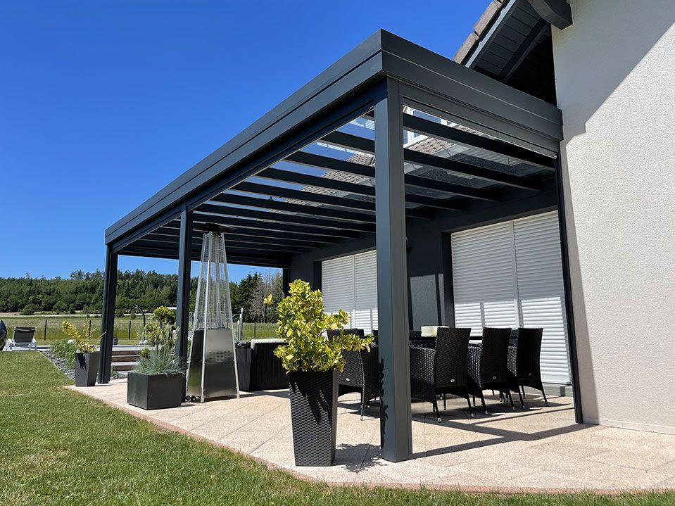 Patio moderne avec pergola noire attenante à une maison blanche, agrémenté de sièges d'extérieur et de plantes en pot au soleil
