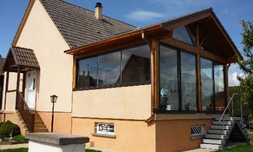 Petite maison beige avec une grande véranda vitrée et un avant-toit en bois.