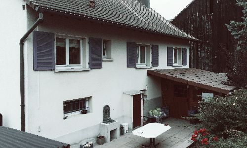 Maison blanche aux volets bleus et patio couvert donnant sur un jardin arboré.