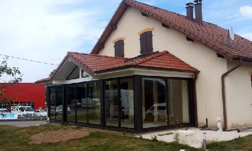 Maison couleur crème avec toit rouge et véranda vitrée dans un jardin gazonné