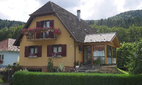 Maison jaune aux volets rouges et balcon fleuri dans un cadre champêtre verdoyant à flanc de colline