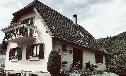 Maison blanche au toit sombre et pentu, volets rouges, jardinières et petit balcon dans un cadre rural