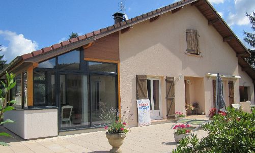 Maison beige de plain-pied avec une véranda vitrée et un patio fleuri sous un ciel bleu.