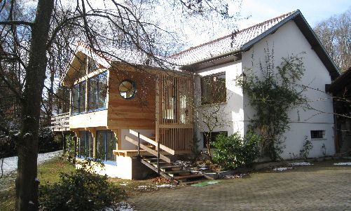 Maison à deux étages avec bardage blanc, balcon en bois et marches d'entrée entourées d'arbres et de verdure.